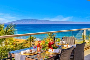 A dining table set with plates, glasses, and a bottle, overlooking a beach and ocean. - Honua Kai | Ocean View 6 BR, Sleeps 20 | Car Incl. w/ 6+ Nights | HKH ML-3101 by KBM (Lahaina)