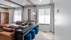 Una habitación moderna con sofá, una mesa de centro y un escritorio con otomanas azules. Hay ventanas con cortinas y un techo con vigas de madera.