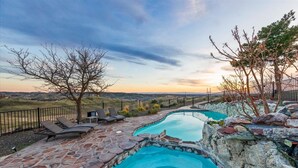 A pool area with stone flooring and two swimming pools, surrounded by a stone wall and a fence.