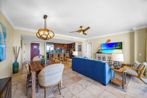 A spacious living area with a dining table, blue sofa, and a TV displaying a beach scene. - 4 Honua Kai Villas | Ocean Front | Sleeps 26 | Free Car w/ 6+ Nights | ML-3113 (Lahaina)