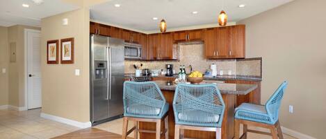 A kitchen with wooden cabinets, a stainless steel refrigerator, a microwave, and a gas stove. There are two blue wicker chairs and a small island with a bowl of fruit.