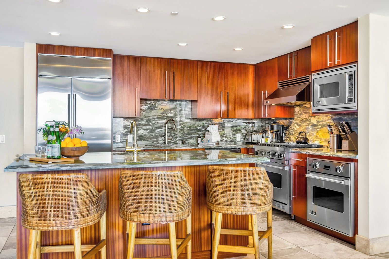 A modern kitchen with wooden cabinets, a large island with bar stools, stainless steel appliances, and a stone backsplash.