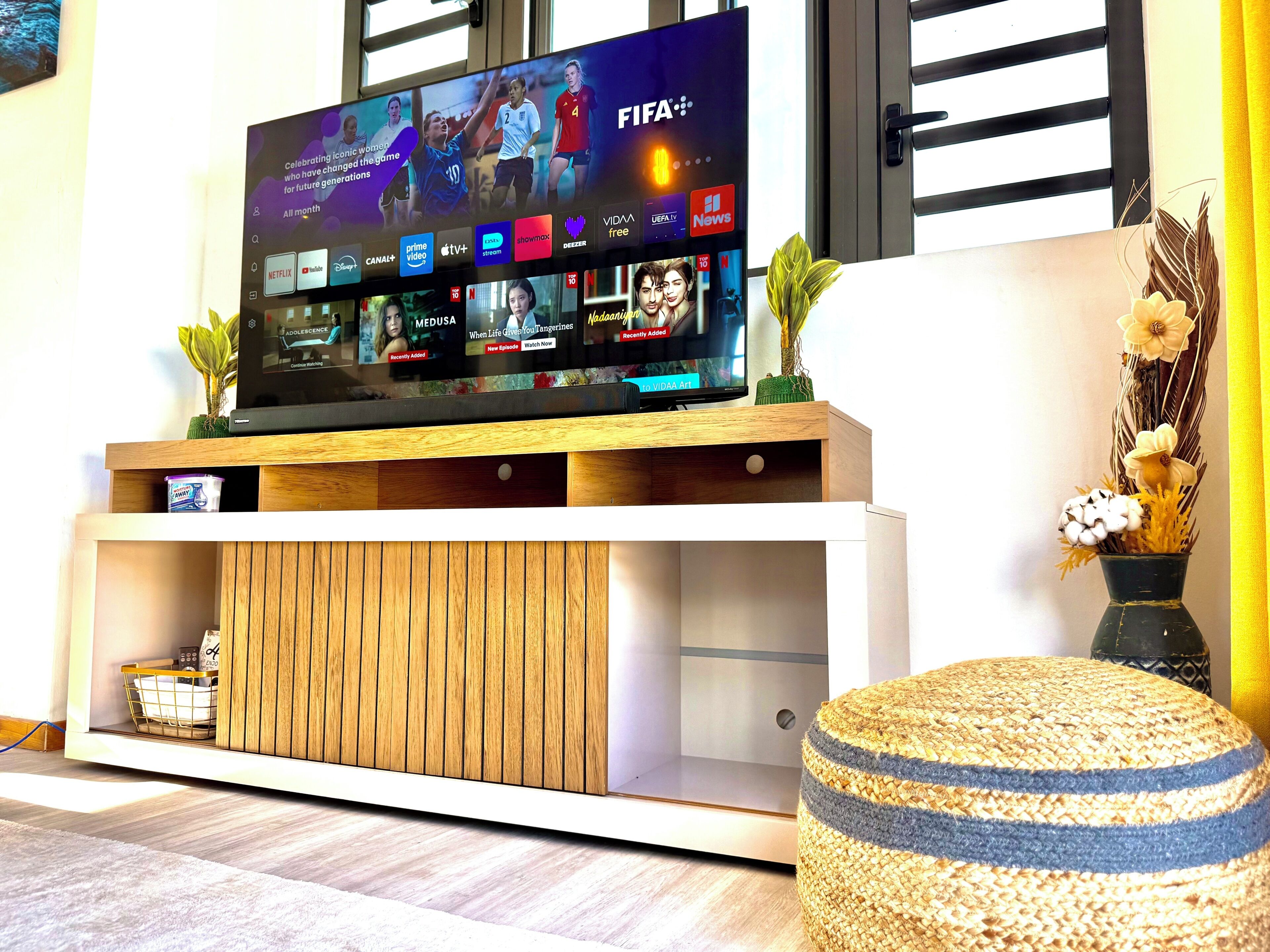 A modern living room with a flat-screen TV displaying various streaming services, a wooden entertainment unit, a woven ottoman, and a decorative vase with dried plants.