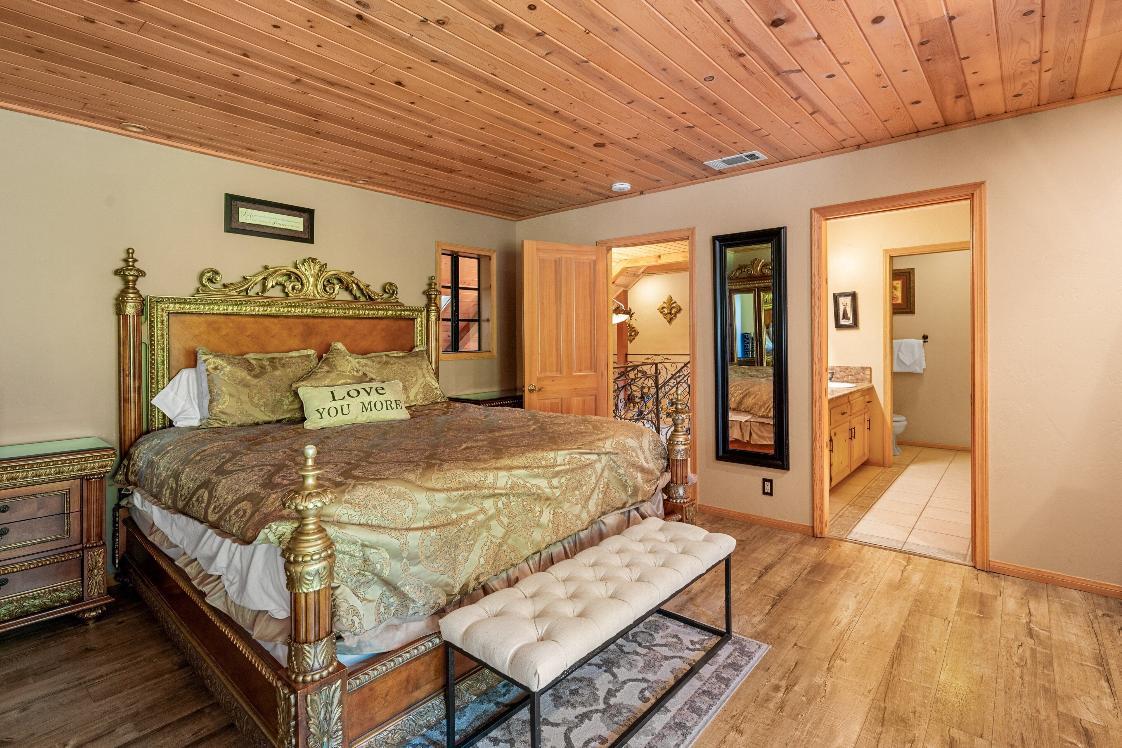 Un dormitorio con una cama grande, un banco, techo de madera y un baño visible a través de una puerta abierta.