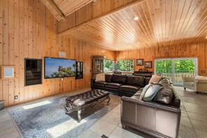 A spacious living room with wooden walls, a leather sofa, a television, and a large window with a view of greenery. - Grand Oriole Estate - Luxury Estate | Pool, Hot Tub, Game Room (Big Bear Lake)