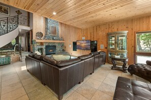 A wooden-paneled room with a stone fireplace, leather sofas, a television, and a glass cabinet. - Grand Oriole Estate - Luxury Estate | Pool, Hot Tub, Game Room (Big Bear Lake)