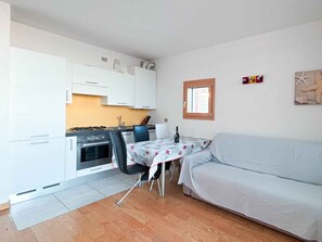A compact kitchen with white cabinets, a gas stove, and a dining area with a table and chairs. - Holiday apartment with balcony (Jesolo)