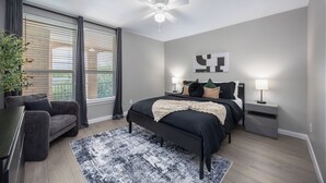 Une chambre à coucher comprenant un lit, des lampes de chevet, un ventilateur de plafond, une grande fenêtre avec des stores et une plante en pot.