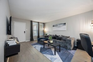 Un salon moderne avec un canapé, une table basse, un téléviseur et une grande fenêtre équipée de stores.