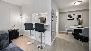 Une chambre moderne comprenant un lit, deux tabourets de bar noirs, une petite table avec un vase, une lampe sur pied et un coin repas avec une table et des chaises blanches.