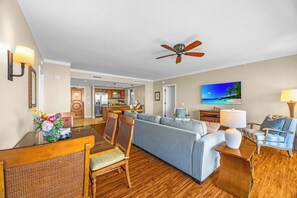 A modern living room with a sofa, a dining table, a TV, and a ceiling fan. - Honua Kai | Ocean View 4 BR, Sleeps 12 | Car Incl. w/ 6+ Nights | HKK ML-3131 by KBM (Lahaina)