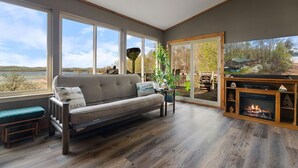 A cozy living room with a sofa, a fireplace, and large windows offering a view of the outdoors. - Lakefront, Private Dock, Water Toys, Sunset Views! (Jefferson City)