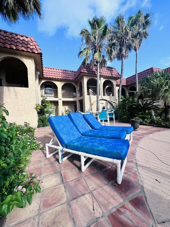 A poolside area with lounge chairs, palm trees, and a building with red-tiled roofs. - Starfish Stay | Tranquil Pool | Directly Across From Beach (Corpus Christi)