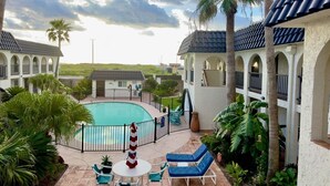 A pool area with lounge chairs and a small table, surrounded by palm trees and a building with a tiled roof. - Starfish Stay | Tranquil Pool | Directly Across From Beach (Corpus Christi)