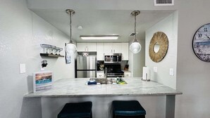 A kitchen with a marble island, two bar stools, stainless steel appliances, pendant lights, and a wall clock. - Starfish Stay | Tranquil Pool | Directly Across From Beach (Corpus Christi)