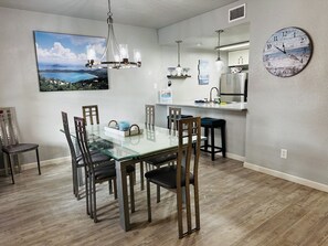 A dining area with a glass table, wooden chairs, a chandelier, a clock, and a kitchen with a stainless steel refrigerator. - Starfish Stay | Tranquil Pool | Directly Across From Beach (Corpus Christi)