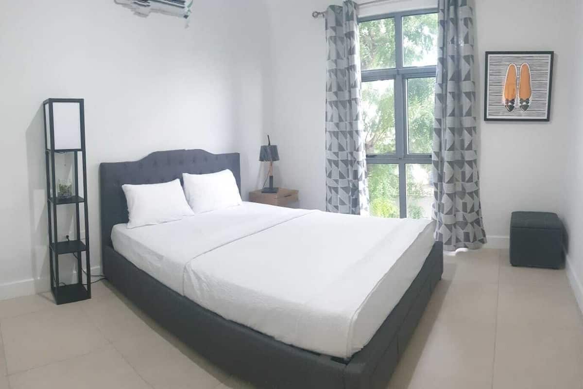 Un dormitorio con una cama, una mesita de noche, una lámpara de pie, una ventana con cortinas y un cuadro en la pared.