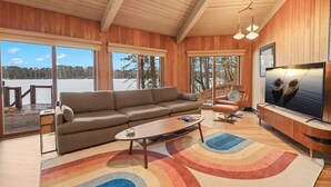 A modern living room with a sofa, armchair, coffee table, and TV. Large windows offer a view of a lake and trees. - Lakefront Retreat with Sandy Beach, Swim Area & Relaxing Lakeside Seating! (Eagle River)