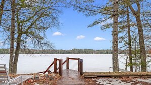 A wooden deck leading to a frozen lake, surrounded by trees. - Lakefront Retreat with Sandy Beach, Swim Area & Relaxing Lakeside Seating! (Eagle River)
