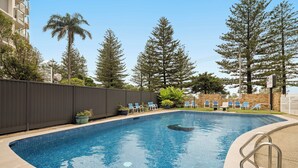 A swimming pool with a tiled edge, surrounded by a fence, and adjacent to a building with a brick wall and a sign. - Coastal Comfort with Ocean Views  (Burleigh Heads)
