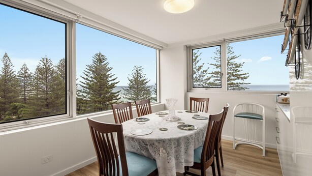 A dining area with a round table, chairs, and a view of trees through large windows. - Coastal Comfort with Ocean Views  (Burleigh Heads)