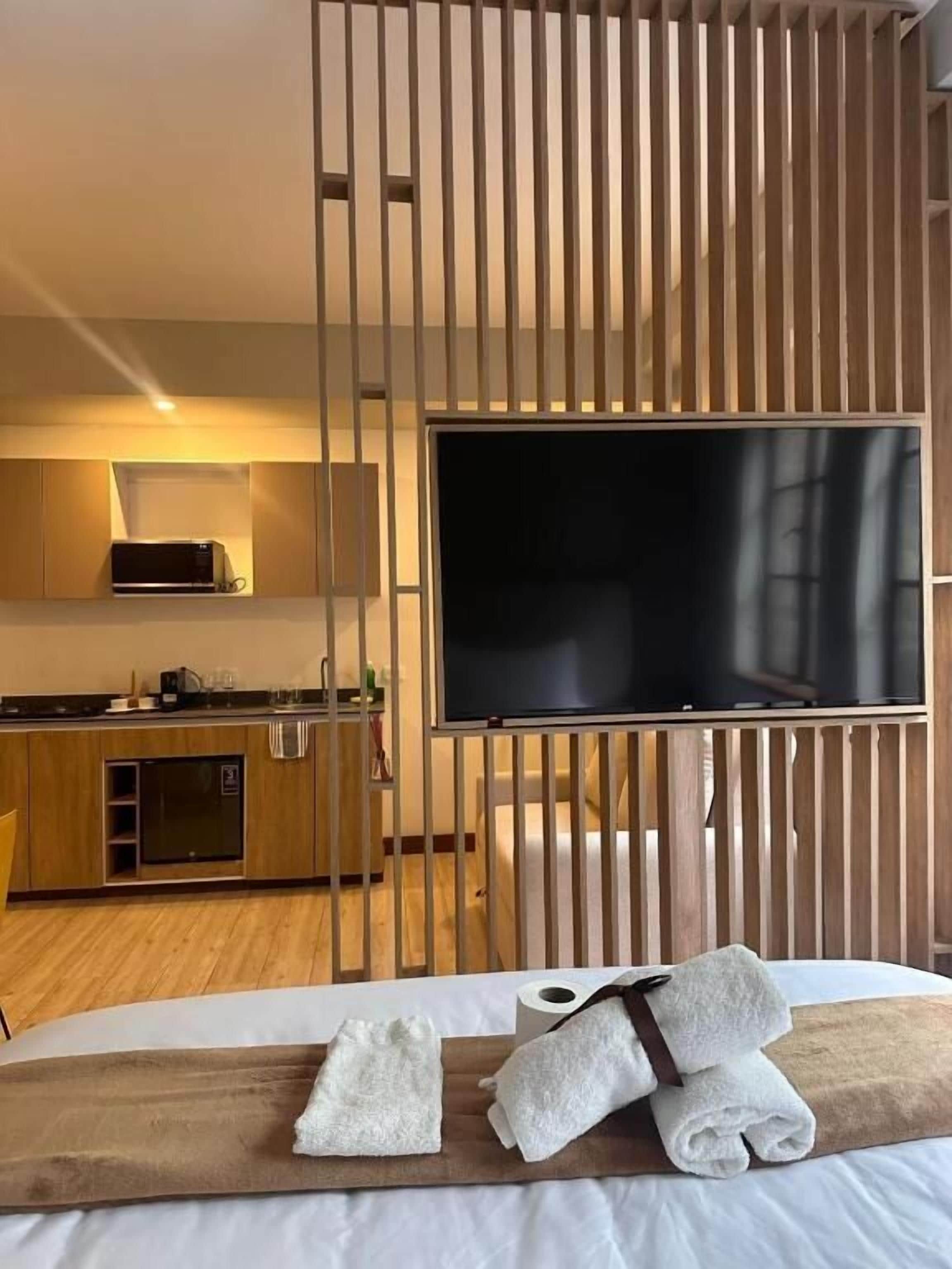 A hotel room with a bed, a TV, a wooden partition, a kitchen area, and towels on the bed.