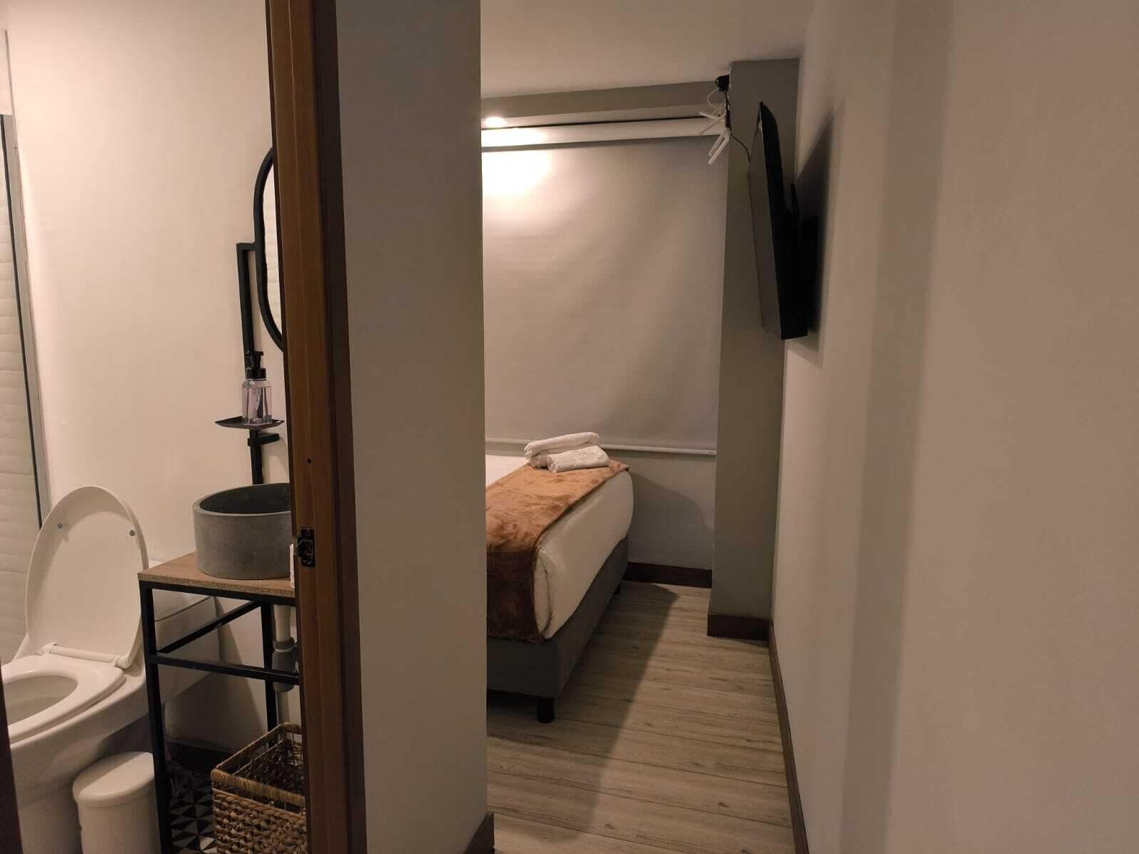 A small, narrow hotel room with a toilet, a bedside table with a bottle, a bed with a blanket, and a TV mounted on the wall.