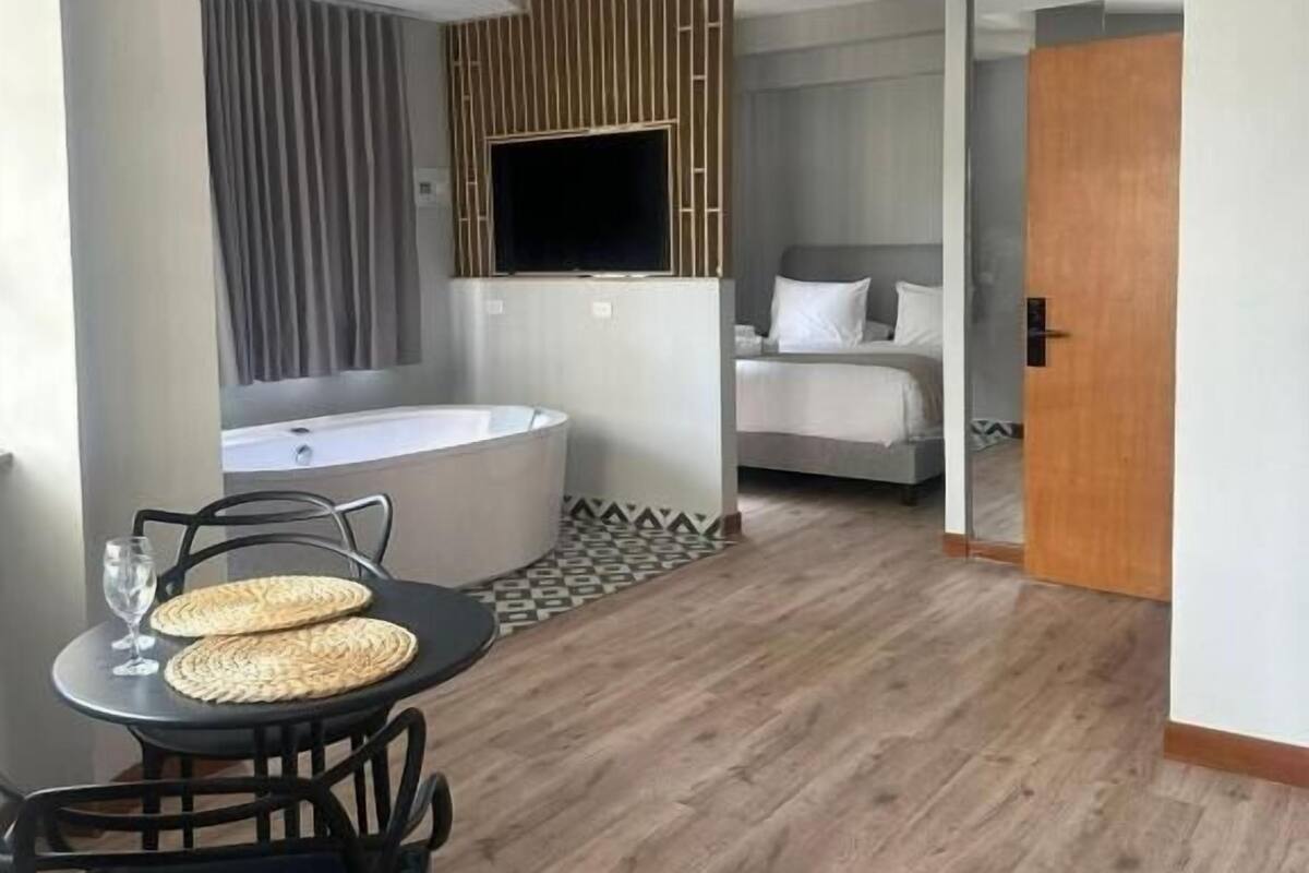 A modern hotel room with a dining area, a large TV, a bathtub, and a bed.