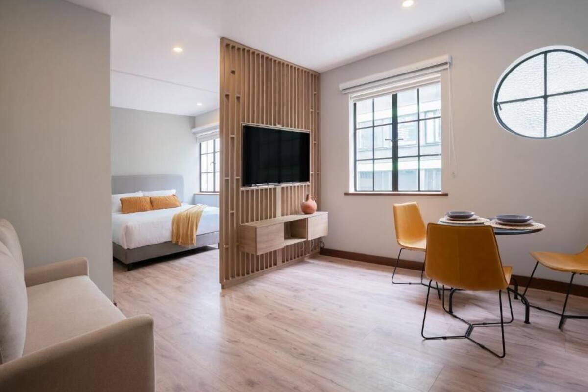 A modern hotel room with a bed, a TV, a dining area with chairs, and a large window.