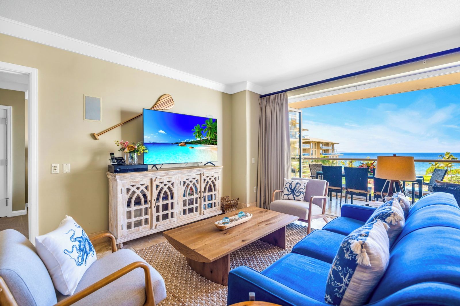Habitación de hotel con televisor grande, mueble de madera, sofá, mesa de centro y balcón con vista al océano.