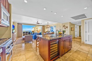 A spacious kitchen with a large island, a dining area with a blue sofa, and a view of a building through the window. - Honua Kai | Ocean View 5 BR, Sleeps 14 | Car Incl. w/ 6+ Nights | HKH ML-3185 by KBM (Lahaina)