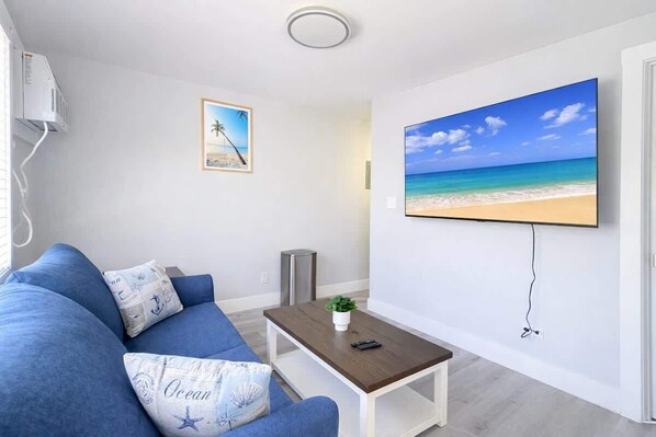 Un salon avec un canapé bleu, une table basse, un tableau représentant une plage et un écran de télévision diffusant une scène de plage.