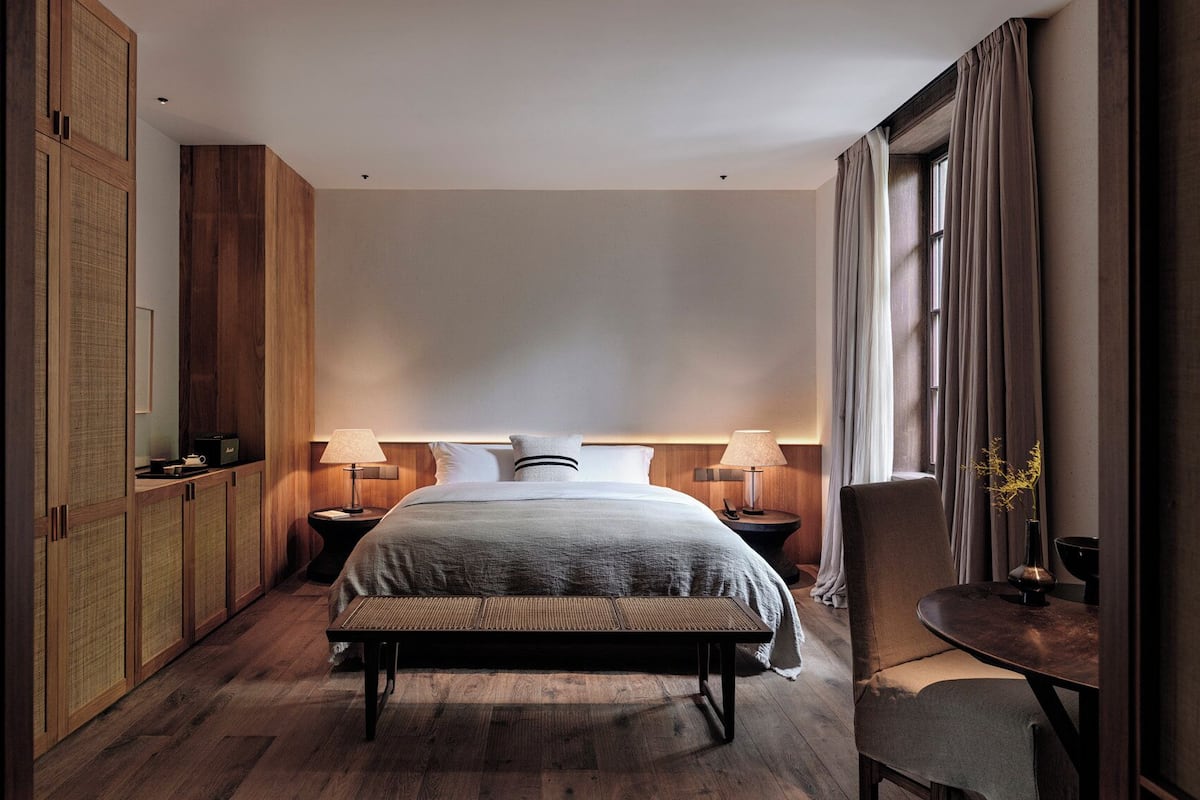 A hotel room with a large bed, two bedside tables with lamps, a bench, a chair, and a large window with curtains.