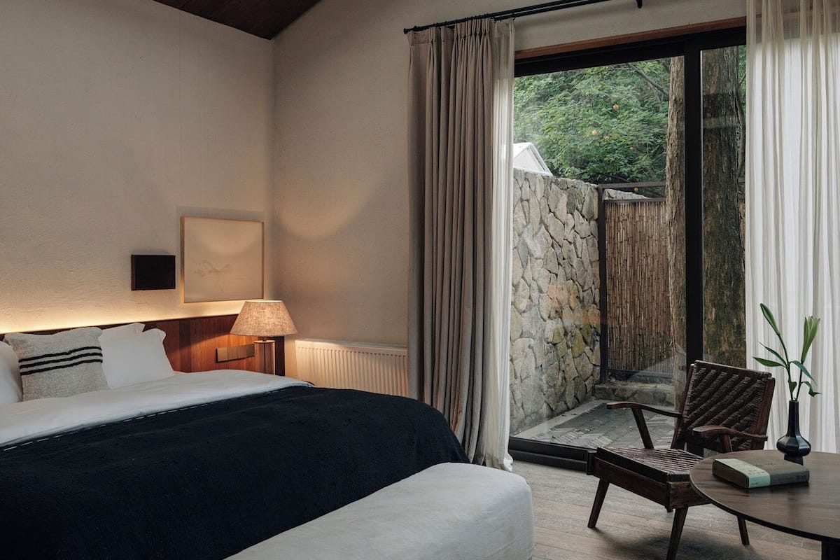 A bedroom with a bed, bedside table, lamp, and a view of a stone wall and greenery outside.