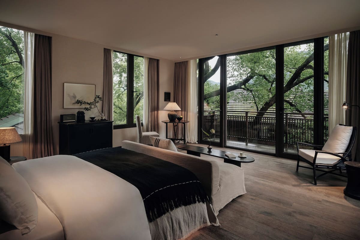 A modern hotel room with a large bed, a desk, a chair, and a balcony with a view of trees.