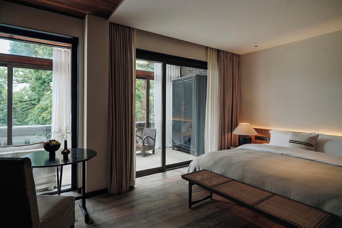 A hotel room with a bed, a small table with a bowl of fruit, a bench, and a view of the outdoors.