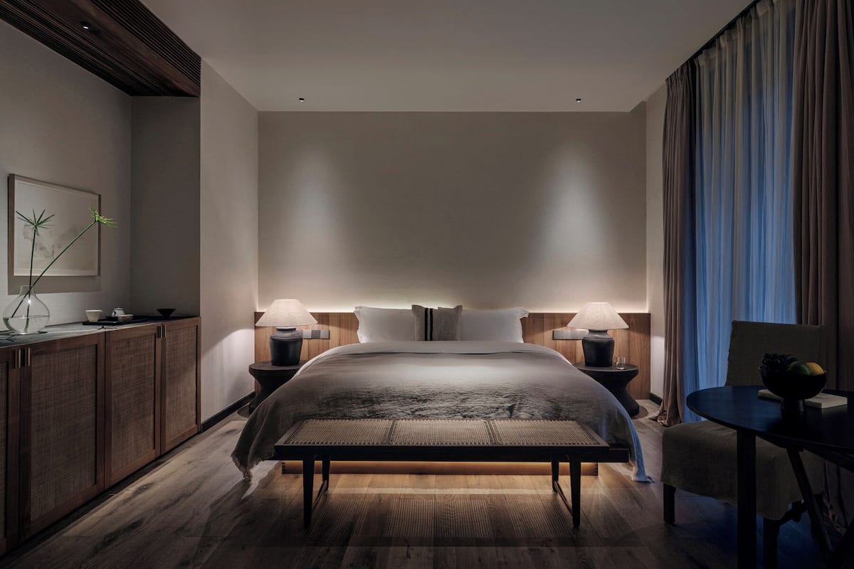 A modern hotel room with a large bed, bedside tables with lamps, a wooden floor, and a view of the city.