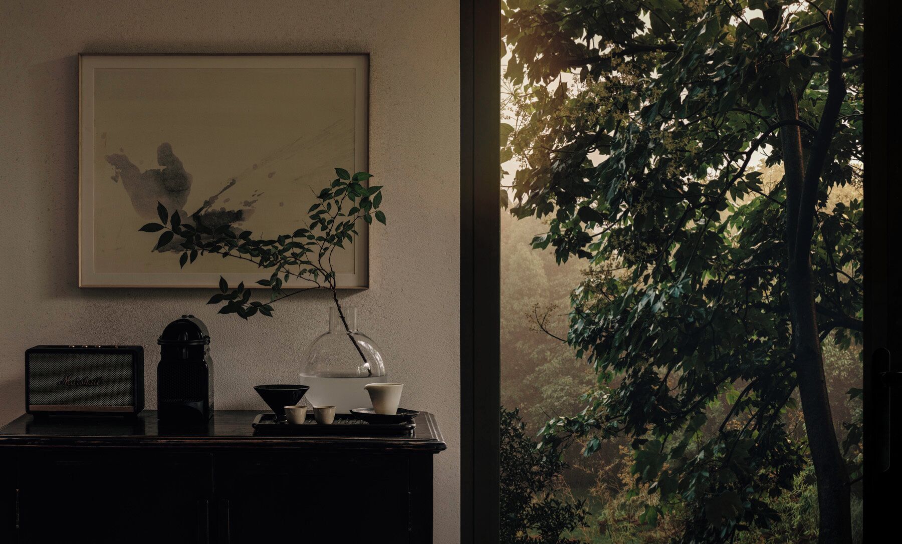 Une œuvre d’art encadrée accrochée au mur, une petite table avec une radio et une fenêtre donnant sur des arbres.