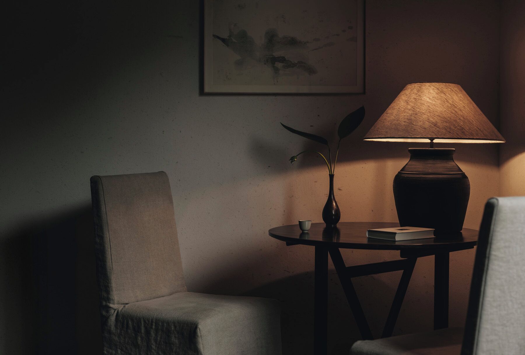A dimly lit room with a table, a chair, a vase with a plant, and a lamp.