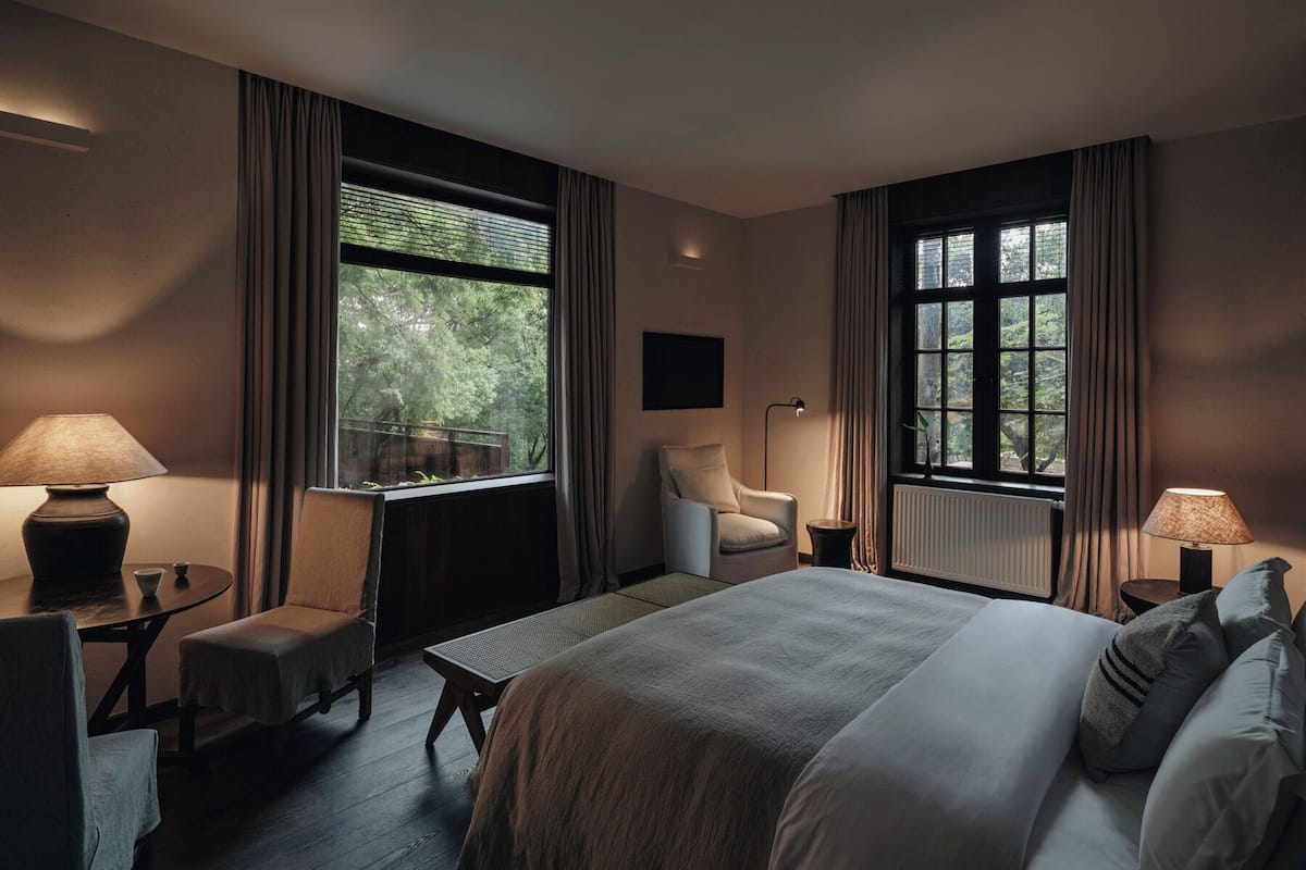 A hotel room with a bed, two chairs, a desk, a lamp, and large windows with curtains.