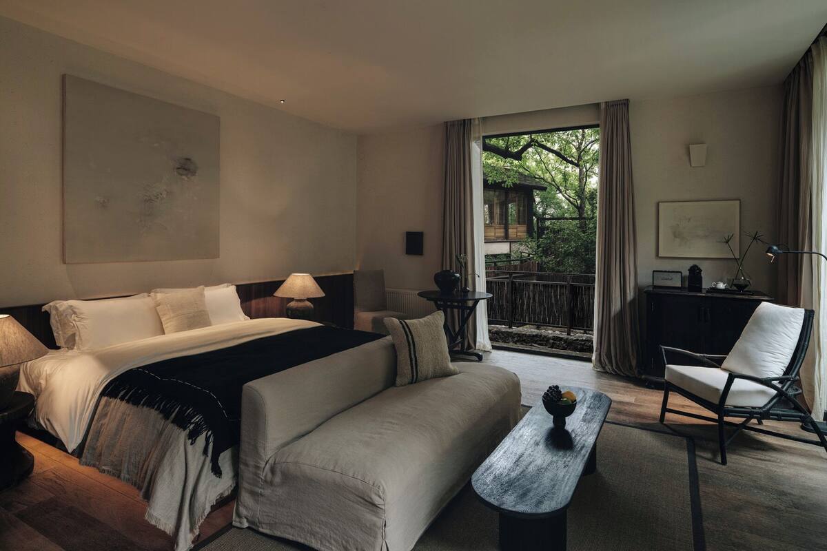 A hotel room with a bed, a sofa, a dining table, a chair, and a balcony with a view of trees.