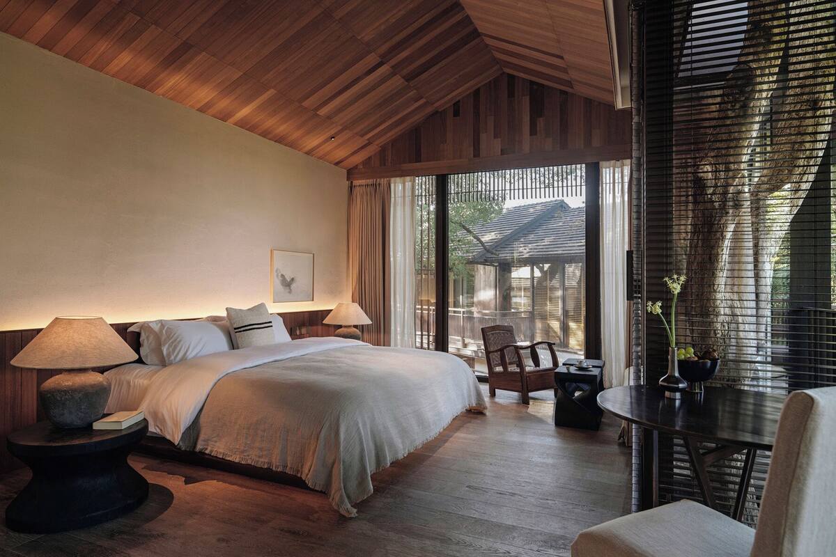 A bedroom with a large bed, bedside lamps, a chair, a small table, and a view of the outdoors.