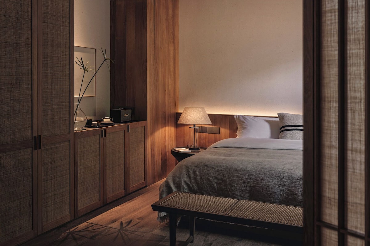 A bedroom with a bed, bedside table, lamp, and a wall-mounted shelf with a plant.