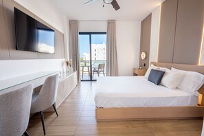 A hotel room with a bed, a desk with two chairs, a TV mounted on the wall, and a balcony with a view of buildings. - Luxury Suite Downtown Punta Cana. 2 min Walk to Coco Bongo • Pool, Gym & Parking (Punta Cana)