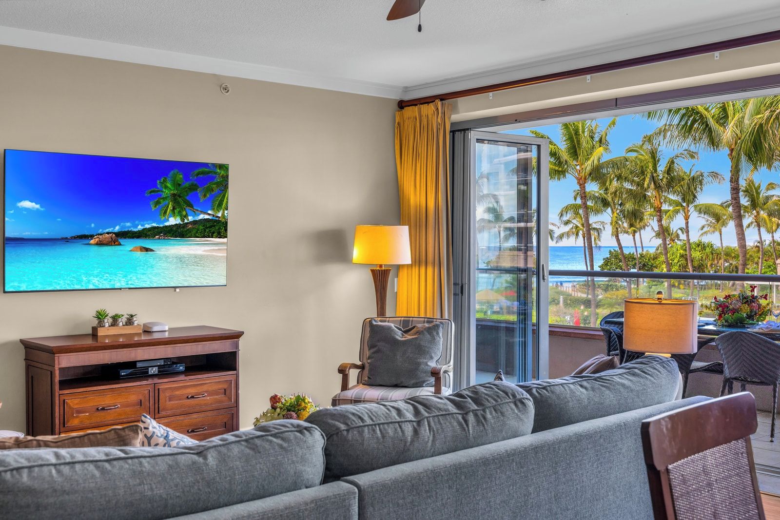 Habitación de hotel con televisor grande, sofá, mesa de comedor con sillas y balcón con vista a palmeras y al océano.