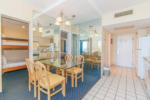 Een kleine hotelkamer met een glazen eettafel, houten stoelen, een stapelbed, een kitchenette en een grote spiegel.