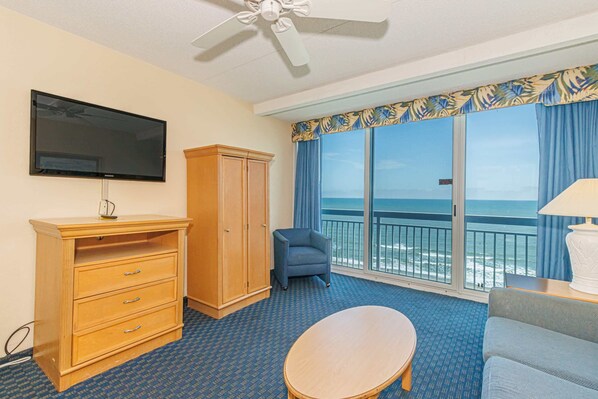 Een hotelkamer met een televisie, een houten dressoir, een blauwe fauteuil, een ronde tafel en een balkon met uitzicht op zee.