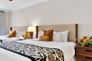 Une chambre d’hôtel avec deux lits, une table de chevet avec une lampe, une commode en bois et un arrangement floral.