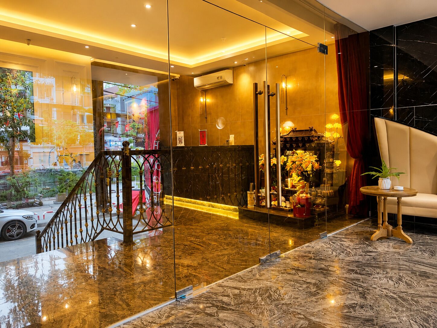 A hotel lobby with a marble floor, a reception desk, a staircase, and a view of the street outside.