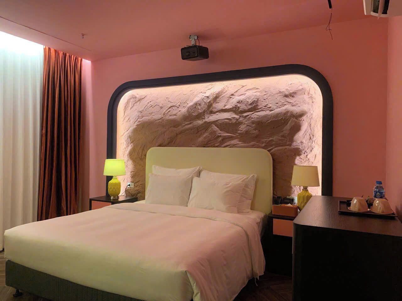 A hotel room with a bed, two bedside tables, a lamp, a water bottle, and a wall-mounted projector.